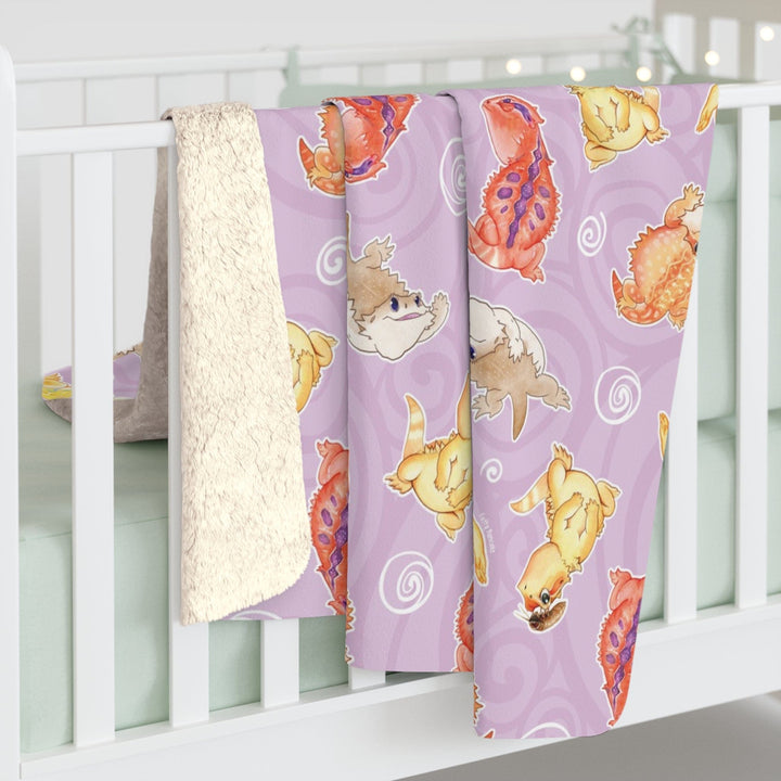 Bearded dragon sherpa blanket with 4 different bearded dragons doing silly things: leaping, giving stink eye, flattening their body, and one with a roach on it's head. The background of the blanket is soft purple with white lineart of swirls.