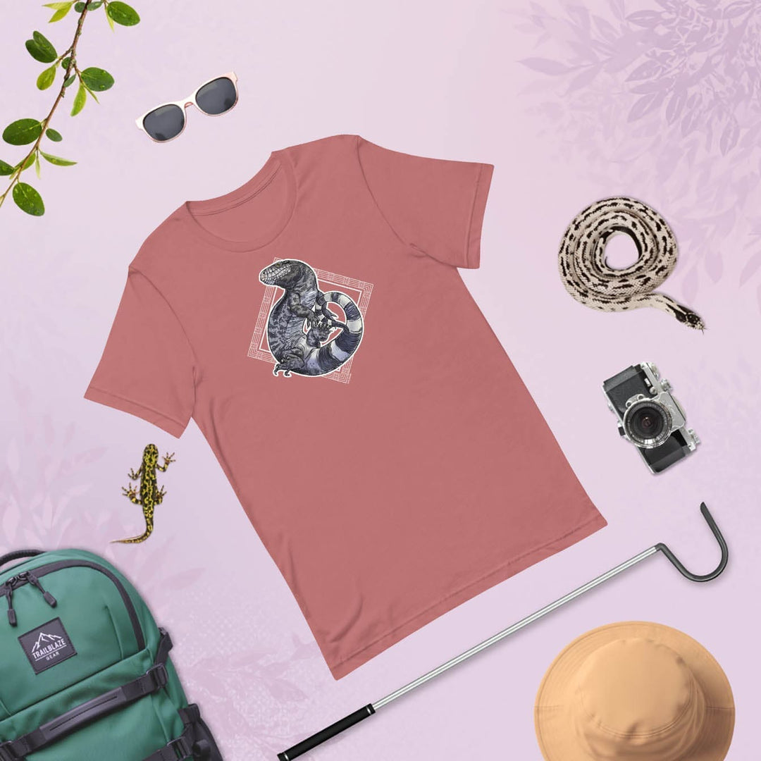 Mauve colored t-shirt flat lay with a tegu lizard surrounded by decorative white elements on a light purple background. It is surrounded by a variety of hiking accessories like a backpack, snake hook, camera, sun glasses, hat with a salamander and snake.