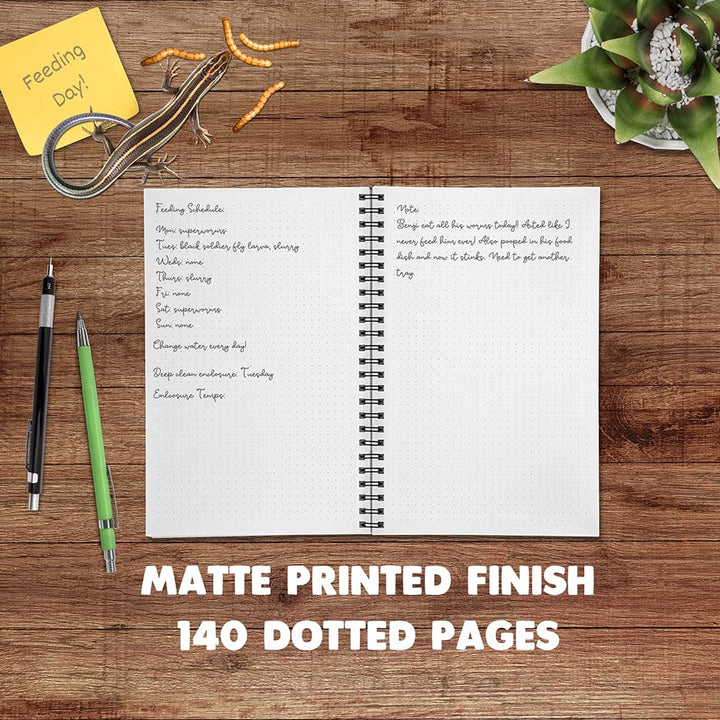 An open spiral-bound planner or notebook with a white matte paper cover spread on a wooden desk, surrounded by pens, a sticky note, a lizard eating worms, and a desk plant. Large text at the bottom reads &