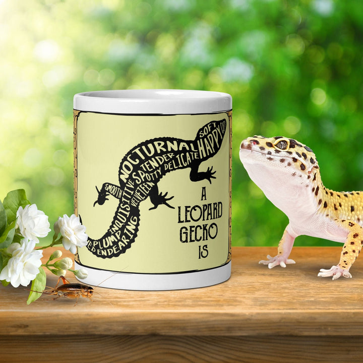 A ceramic mug with a yellow and black design of a leopard gecko sitting on a wooden table top with a natural background. A real leopard gecko lizard stands next to the mug and white flowers sit on the table on the left.