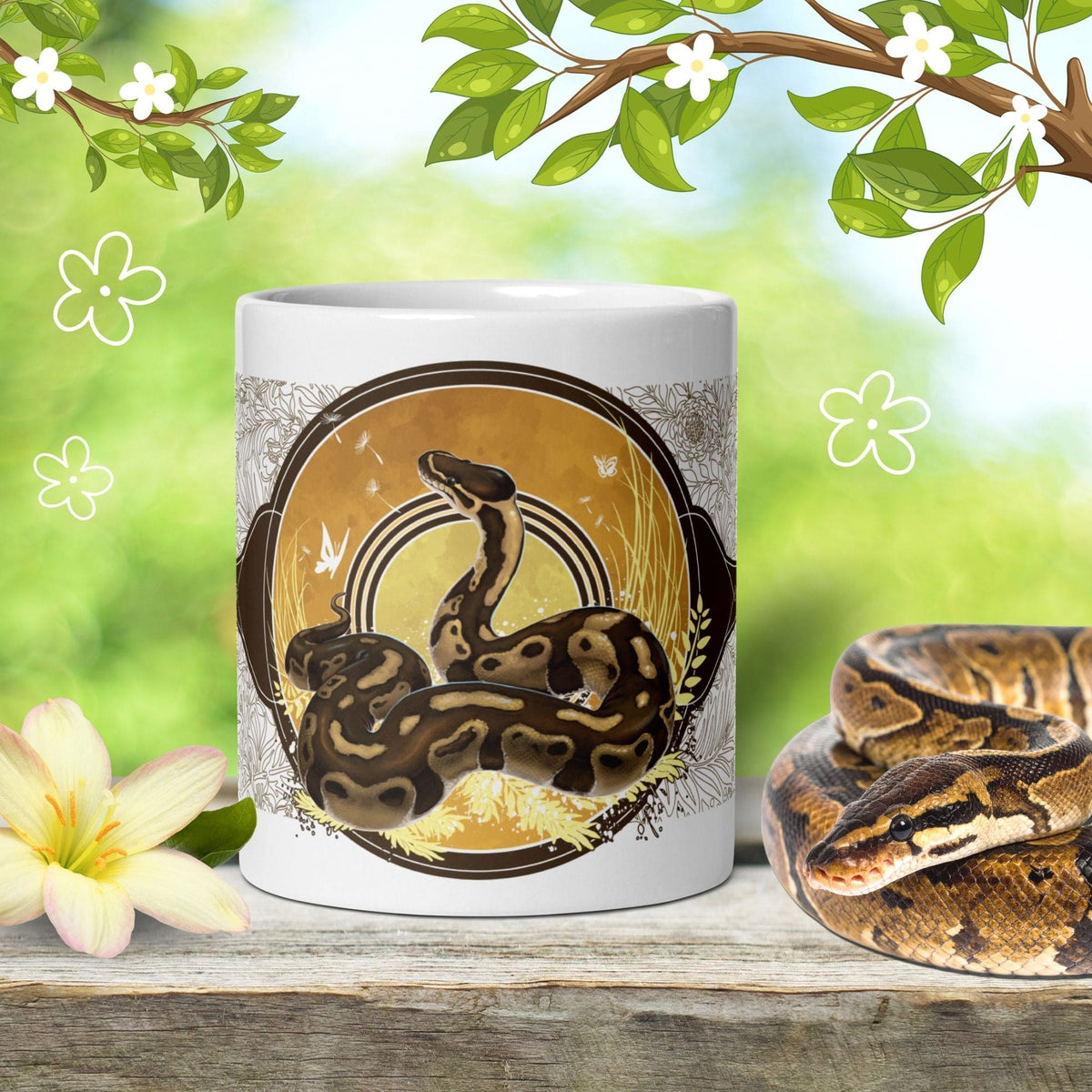 A ceramic mug with a decorative graphic of a brown ball python framed by a circle with gold coloration and yellow grasses. A real ball python sits next to the mug on a wood table with cartoon leaves and flowers surrounding it.