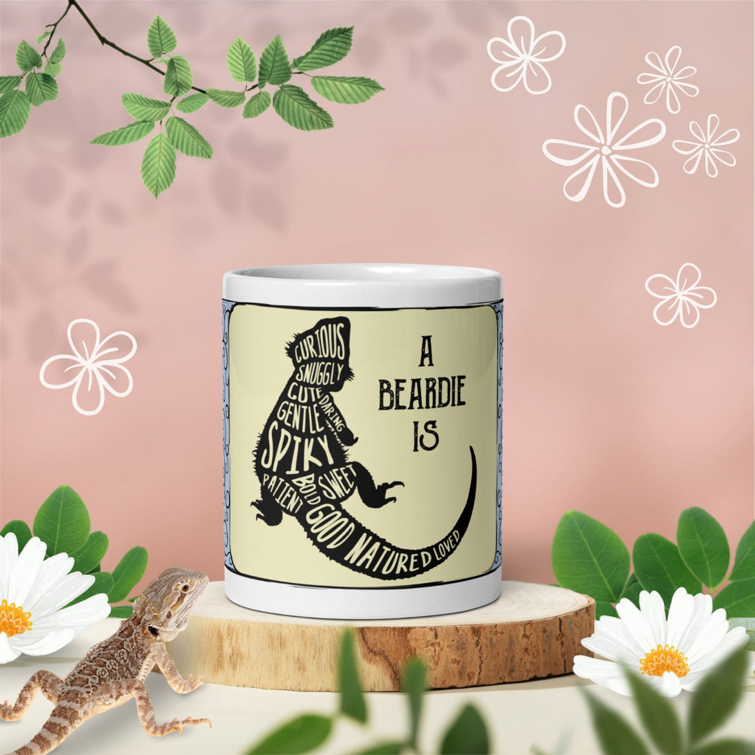  A white ceramic mug featuring a black silhouette of a bearded dragon filled with descriptive words including curious, snuggly, cute, daring, gentle, spiky, sweet, patient, bold, good natured, and loved, with the text A BEARDIE IS.