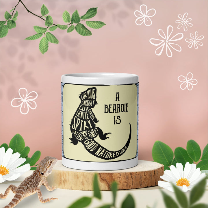  A white ceramic mug featuring a black silhouette of a bearded dragon filled with descriptive words including curious, snuggly, cute, daring, gentle, spiky, sweet, patient, bold, good natured, and loved, with the text A BEARDIE IS.