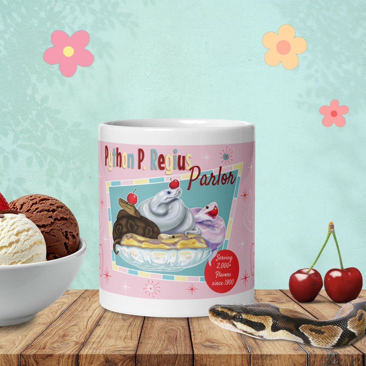 A white ceramic mug featuring a graphic of three ball python snakes in an ice cream glass dish to look like a banana split sundae. The text reads Python P. Regius Parlor and Serving 2,000+ Flavors since 1900. The design includes red cherries, starburst accents, and a pink background.