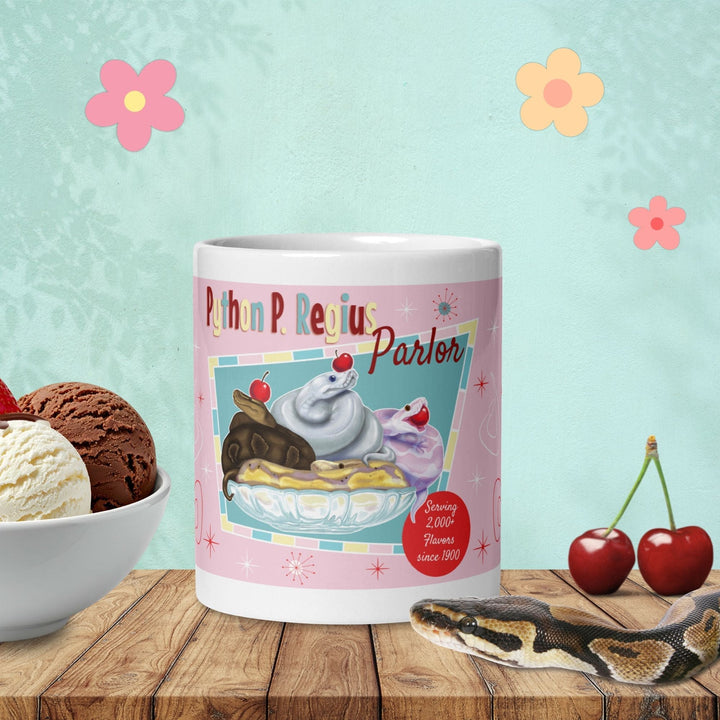 A white ceramic mug featuring a graphic of three ball python snakes in an ice cream glass dish to look like a banana split sundae. The text reads Python P. Regius Parlor and Serving 2,000+ Flavors since 1900. The design includes red cherries, starburst accents, and a pink background.