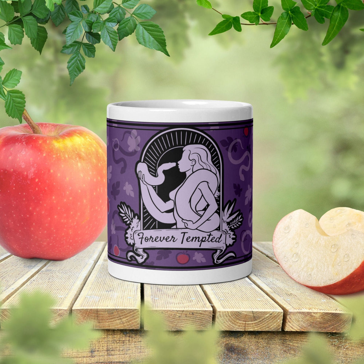 Purple mug with silhouette of woman holding and facing a snake with the text below it reading "Forever Tempted". It sits on a wooden table with apples on either side and a lush green background behind it.