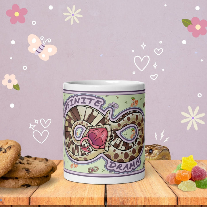A white ceramic mug featuring a graphic illustration of a brown and tan patterned snake coiled into an infinity symbol shape with its mouth open, accompanied by the text "INFINITE DRAMA" in a purple font against a light green background.
