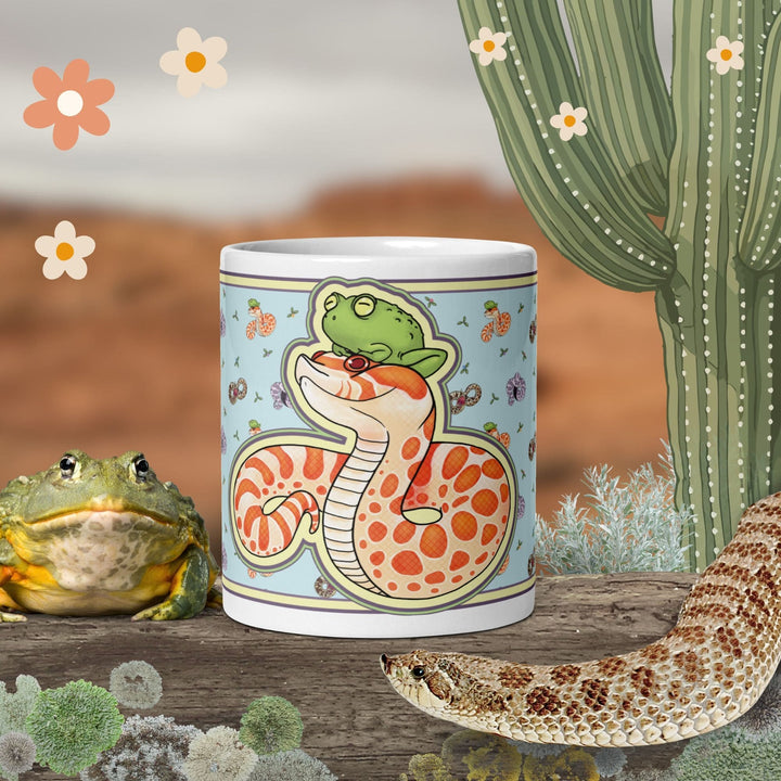 A ceramic mug sitting on a wood log with graphical assets surrounding it. The mug has a graphic of a cartoon albino hognose snake with a frog sitting on it&