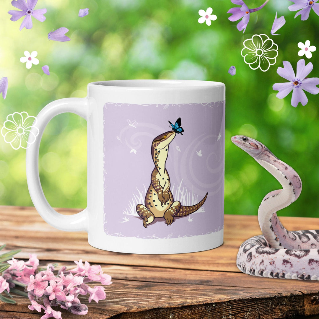 A white ceramic mug featuring a graphic illustration of a brown spotted lizard sitting upright with a small blue butterfly perched on its nose, set against a light purple background with subtle swirling patterns and white grass. It rests on a table with a green background and graphic flowers around it. 