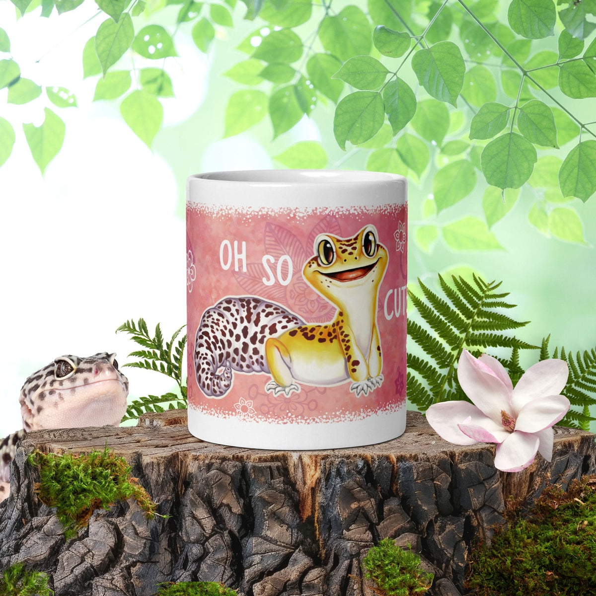Image depicts a white ceramic mug with a graphic of a leopard gecko on it. There are flowers around the cute smiling gecko and text that reads "Oh So Cute". The mug rests on a mossy wooden stump with a real leopard gecko peeking over the edge of the wood.