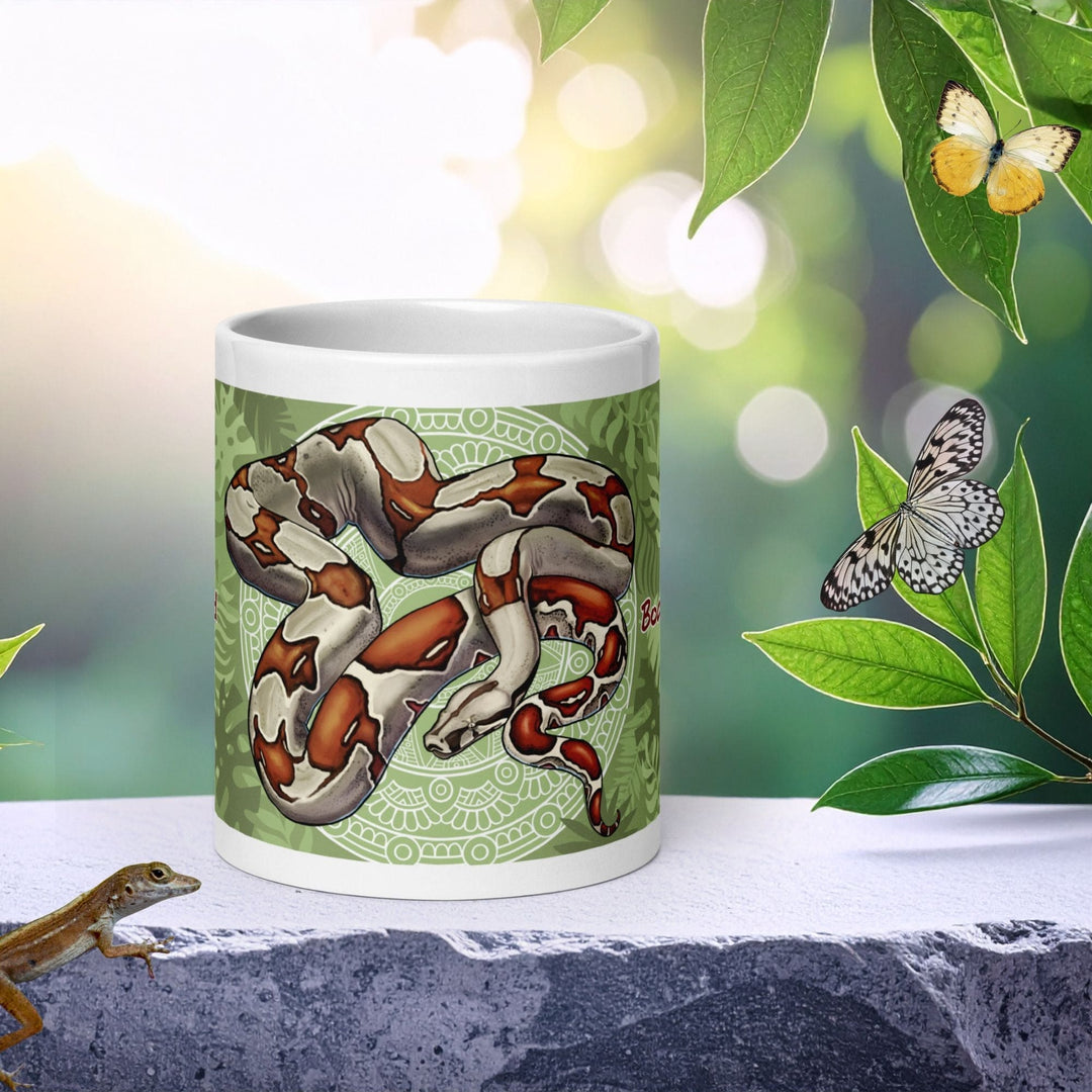 A white ceramic mug featuring a detailed illustration of a green and brown red tailed boa constrictor snake coiled on a decorative circular background, set on a stone ledge with a lizard and butterflies in a blurred garden background.