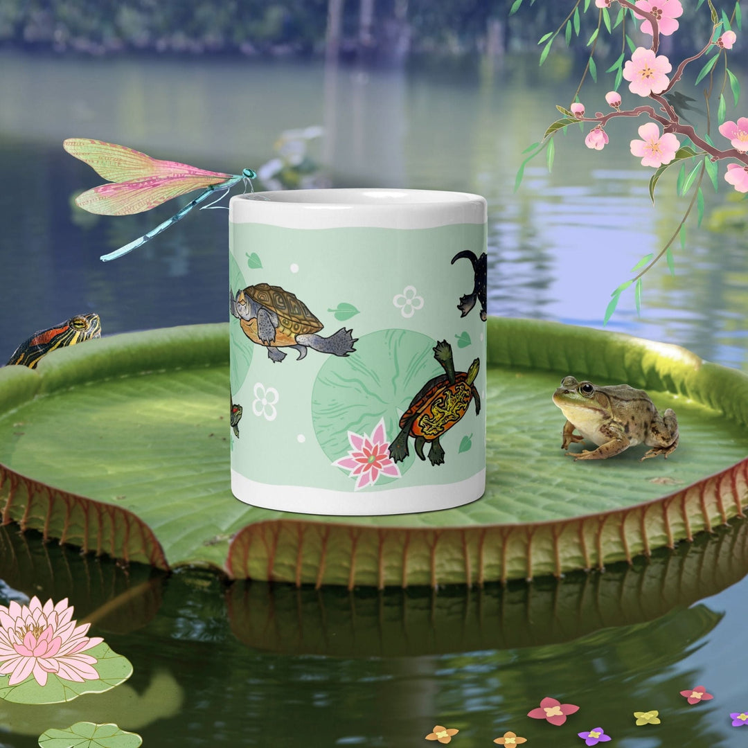A white ceramic mug featuring a light green background with a repeating pattern of small turtles, pink lotus flowers, and green lily pads. The design includes stylized white floral accents scattered across the surface of the mug.