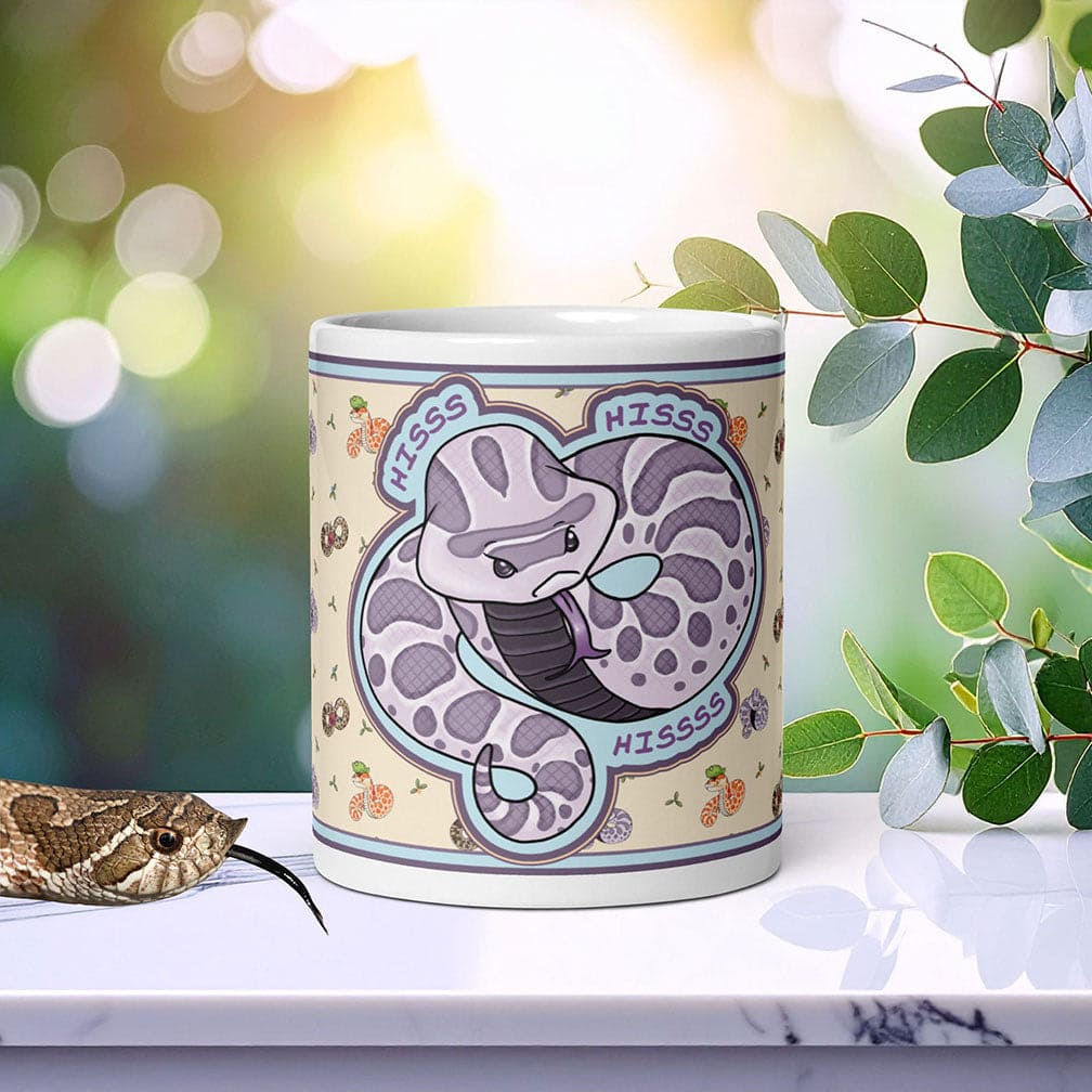 A white ceramic mug with a purple and gray snake design. The mug has the word 'HISS' written on it three times. The mug is sitting on a white marble slab with a real hognose slithering next to the mug.