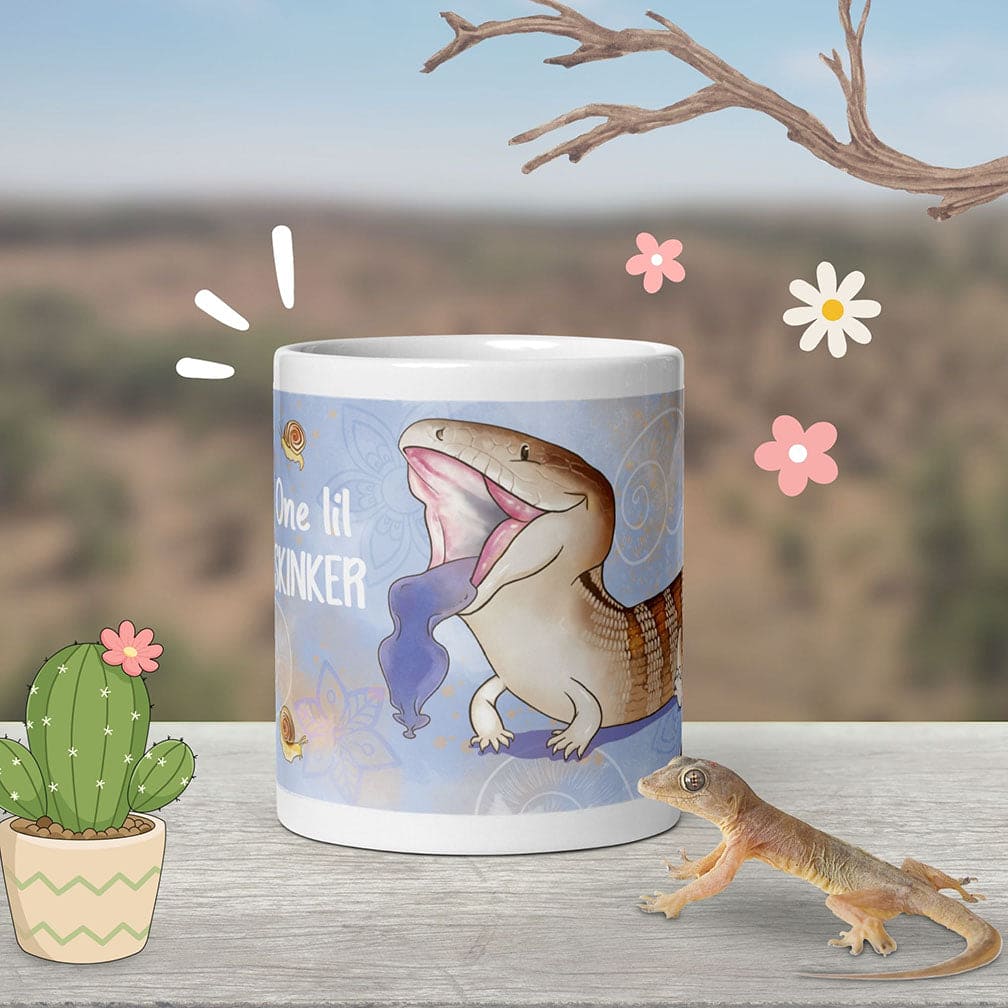 A white ceramic mug with a blue wrapped design featuring a cartoony blue tongue skink lizard with an open mouth, extending out it's tongue, with flowers in the background, and the white text reading "One lil SKINKER" next to it. It sits on a wooden table with a semi-arid background and graphic flowers surrounding it.