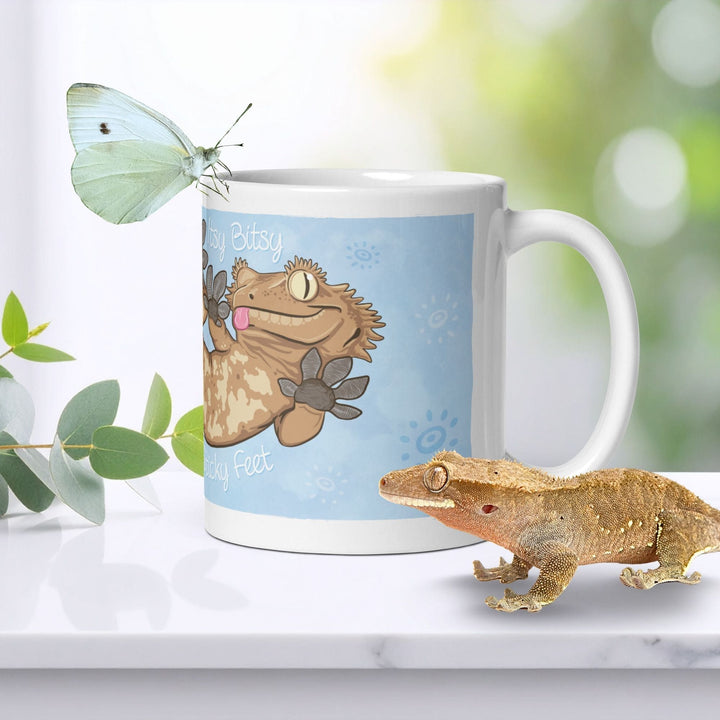  A white ceramic mug featuring a cartoon illustration of a tan crested gecko with the text "Itsy Bitsy Sticky Feet" on a light blue background, accompanied by a real tan crested gecko and a white butterfly perched on the rim.