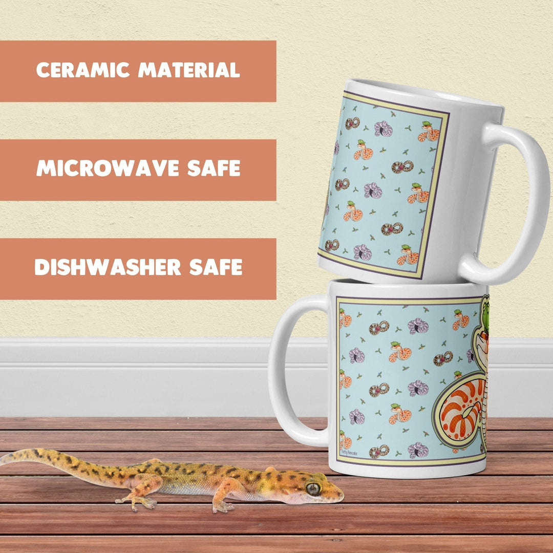 Two white ceramic mugs featuring a light blue panel with a repeating pattern of small colorful snakes. The text on the image reads CERAMIC MATERIAL, MICROWAVE SAFE, and DISHWASHER SAFE. A small brown and yellow spotted lizard is in the foreground.