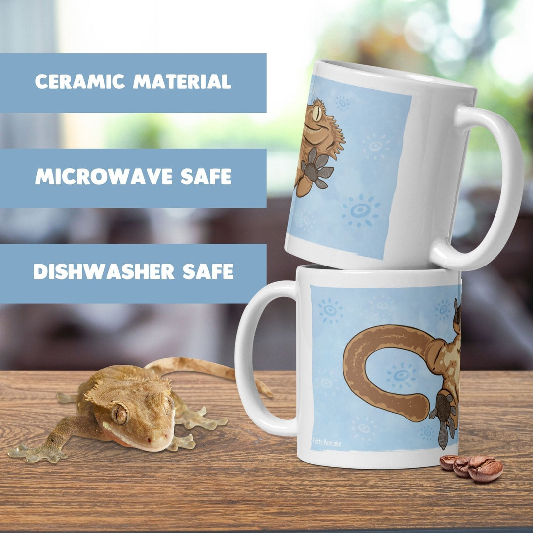 Two white ceramic mugs featuring a cartoon illustration of a crested gecko on a light blue background with sun motifs. The text on the image reads CERAMIC MATERIAL, MICROWAVE SAFE, and DISHWASHER SAFE. A live crested gecko is nearby.