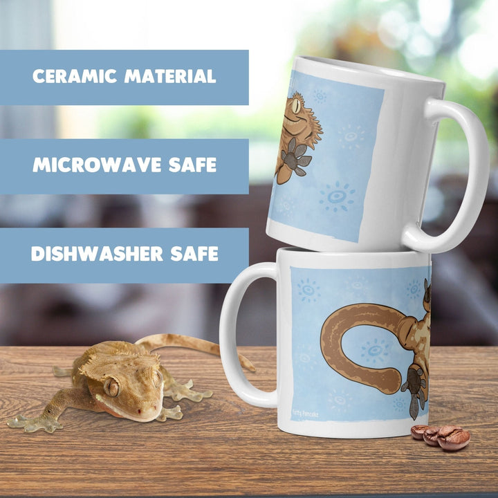 Two white ceramic mugs featuring a cartoon illustration of a crested gecko on a light blue background with sun motifs. The text on the image reads CERAMIC MATERIAL, MICROWAVE SAFE, and DISHWASHER SAFE. A live crested gecko is nearby.