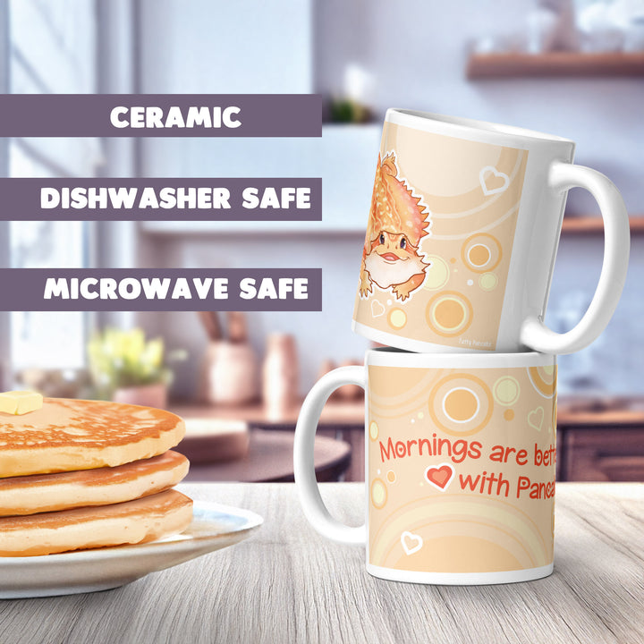 Two white ceramic mugs featuring a cartoon bearded dragon illustration, light orange circular patterns, and hearts. The text reads, "Mornings are better with Pancakes." The mugs are labeled as ceramic, dishwasher safe, and microwave safe.