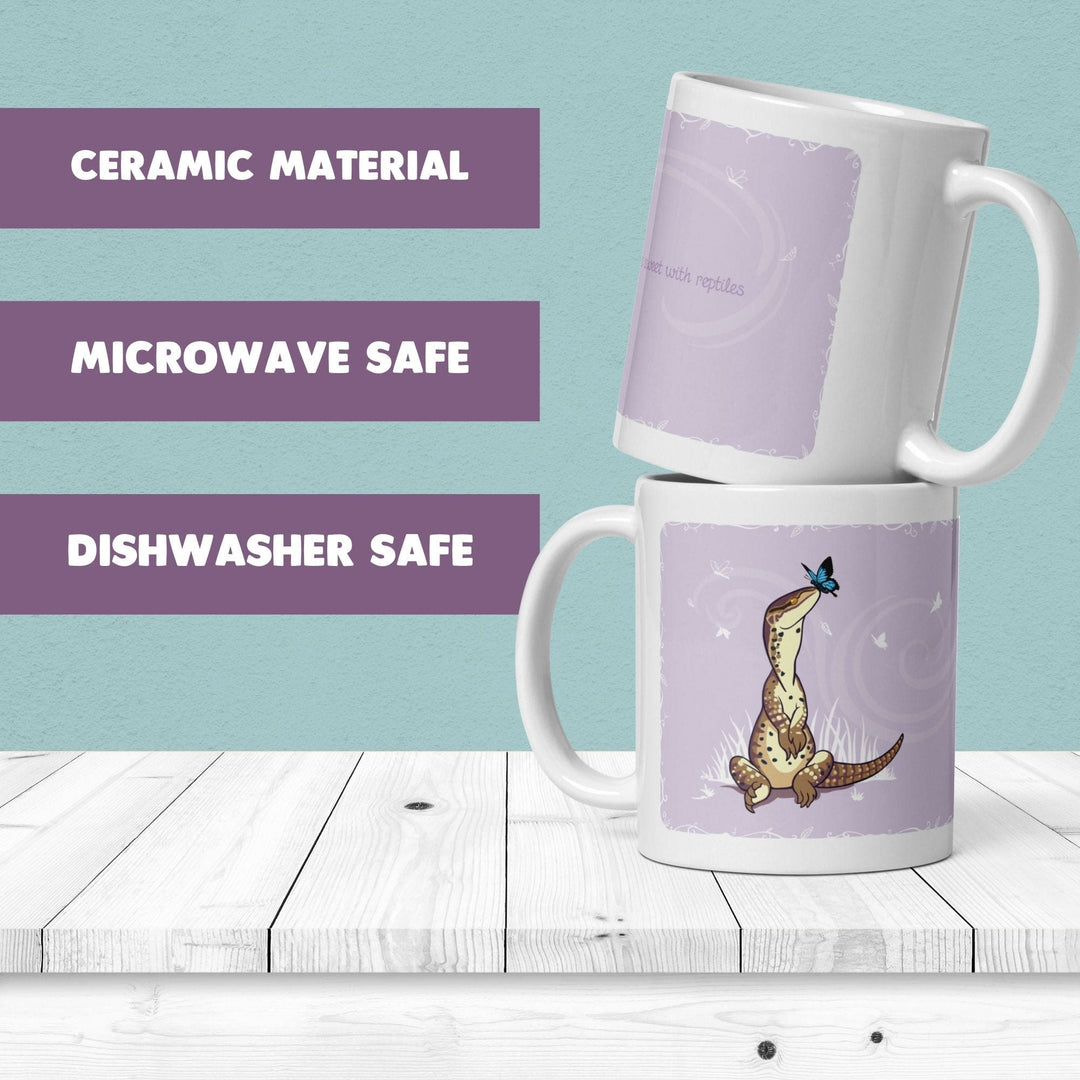 Two stacked white ceramic mugs with a lavender illustration of a cartoon lizard on the front, shown on a white wooden surface against a light blue background. Purple text blocks on the left read &