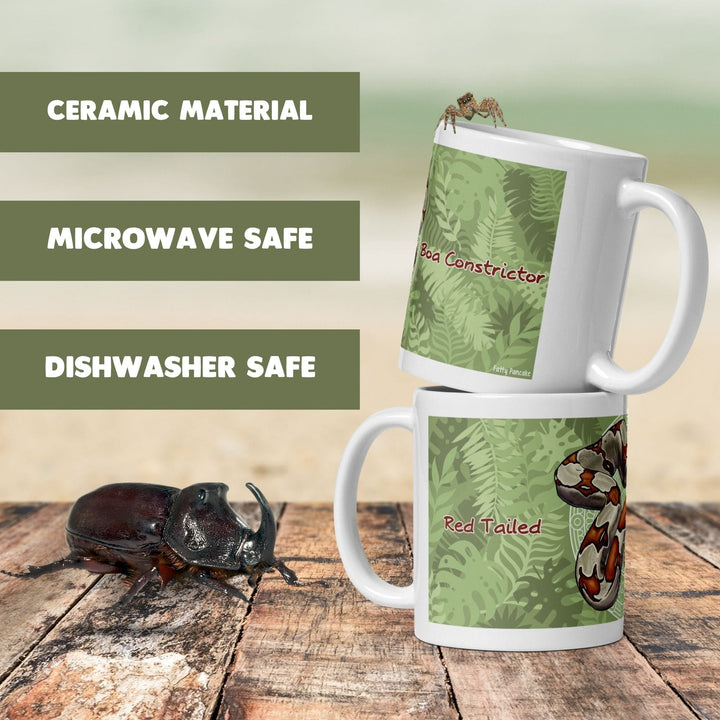 Two white ceramic mugs stacked vertically, featuring a green jungle leaf pattern, a red-tailed boa constrictor illustration, and the text "Boa Constrictor" and "Red Tailed". A dark brown rhinoceros beetle is positioned on the wooden surface. Text overlays on the left read &