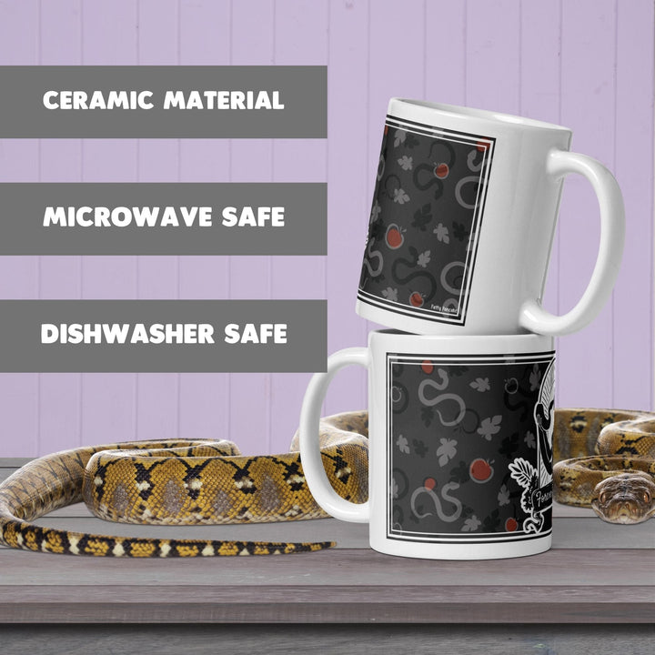 Two white ceramic mugs stacked vertically, featuring a dark gray pattern of snakes, leaves, and red apples. The text on the left reads CERAMIC MATERIAL, MICROWAVE SAFE, and DISHWASHER SAFE. A yellow and brown patterned snake is coiled below.