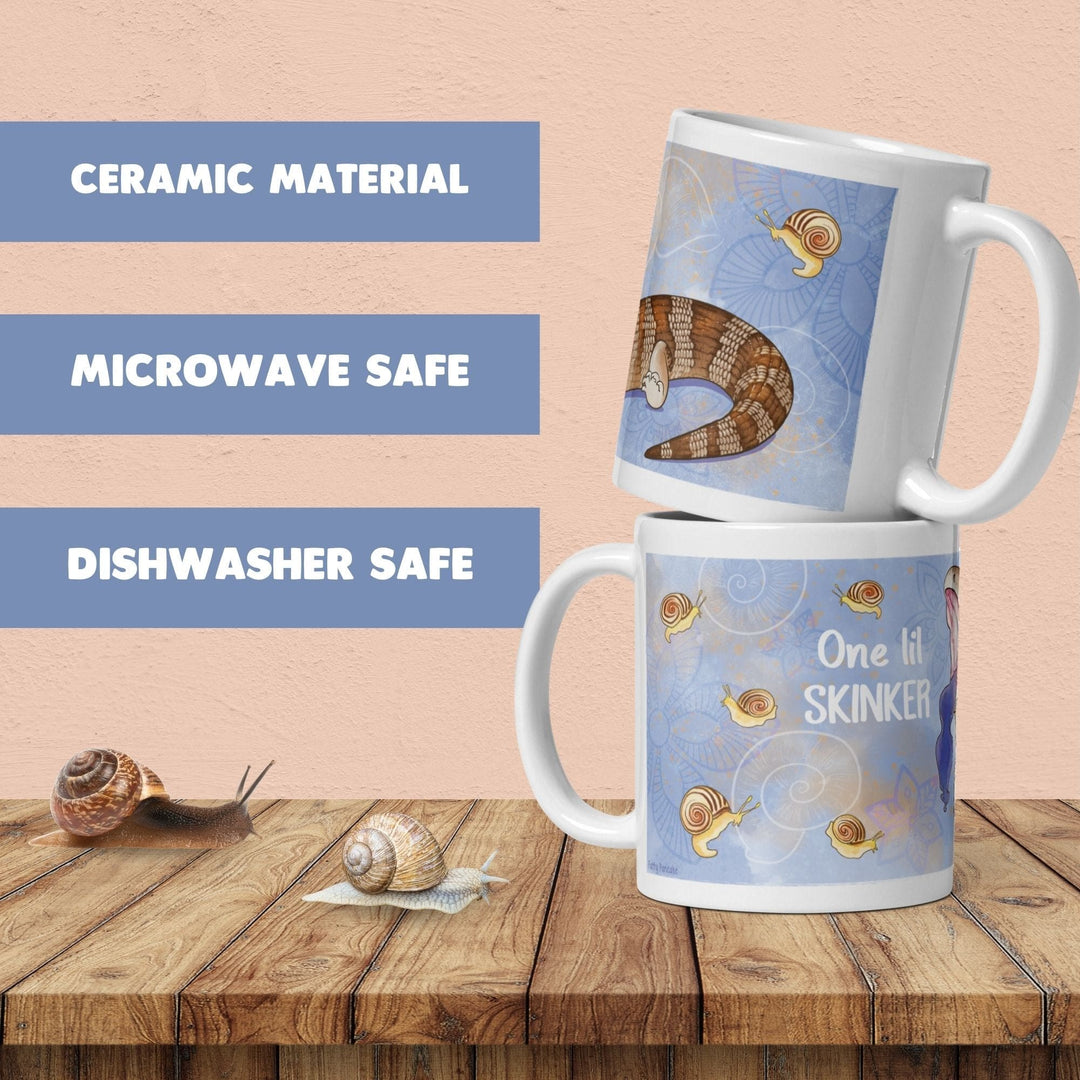 Two white ceramic mugs featuring a blue background with illustrations of snails and a lizard tail. The text on the mugs reads One lil SKINKER. The mugs are microwave and dishwasher safe, as indicated by the text on the left side.