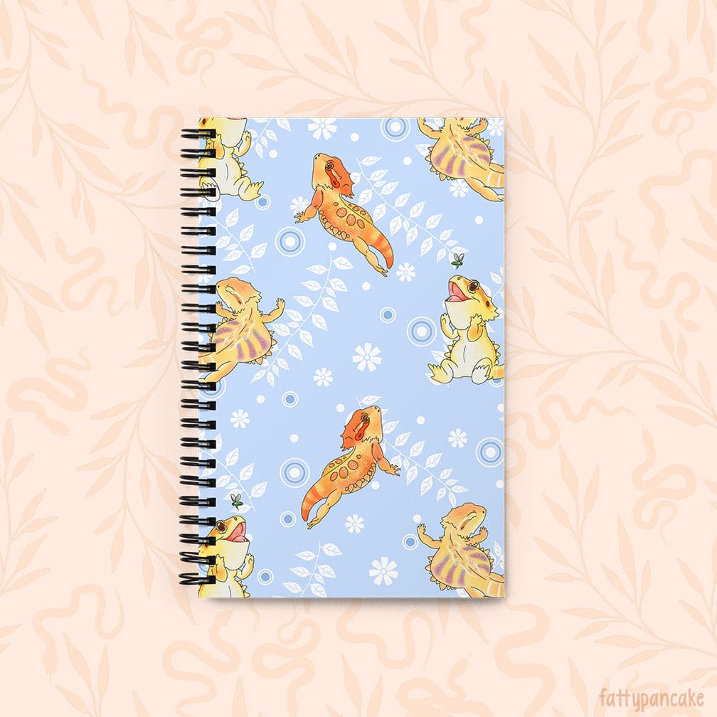 A spiral-bound notebook with a light blue cover decorated with cartoon bearded dragons in yellow and orange with white vines with leaves and circles, set against a pale peach background with a subtle leaf pattern.