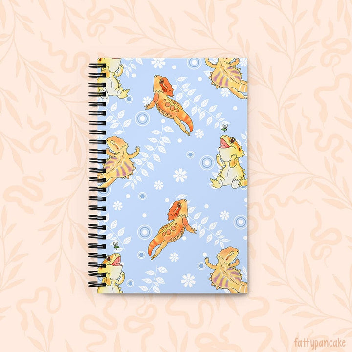 A spiral-bound notebook with a light blue cover decorated with cartoon bearded dragons in yellow and orange with white vines with leaves and circles, set against a pale peach background with a subtle leaf pattern.