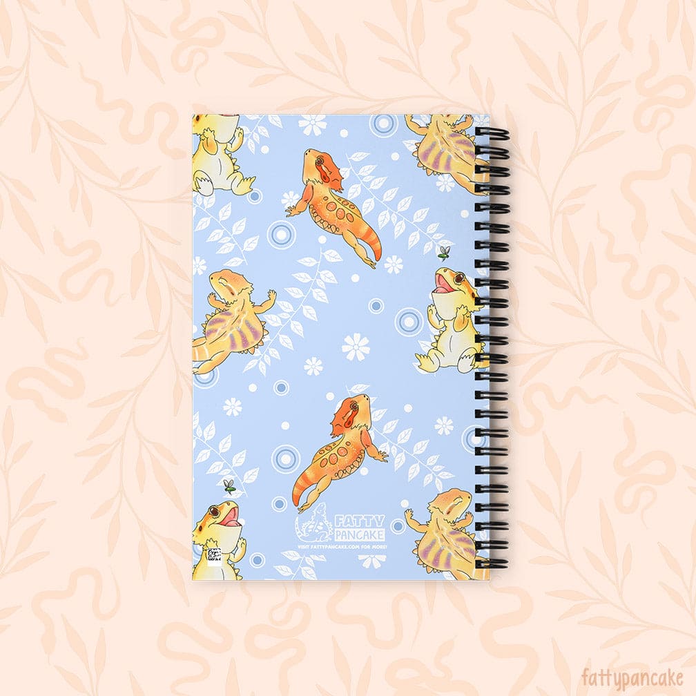 A spiral-bound notebook with a light blue cover decorated with cartoon bearded dragons in yellow and orange with white vines with leaves and circles, set against a pale peach background with a subtle leaf pattern. The logo with Fatty Pancake is at the center bottom.