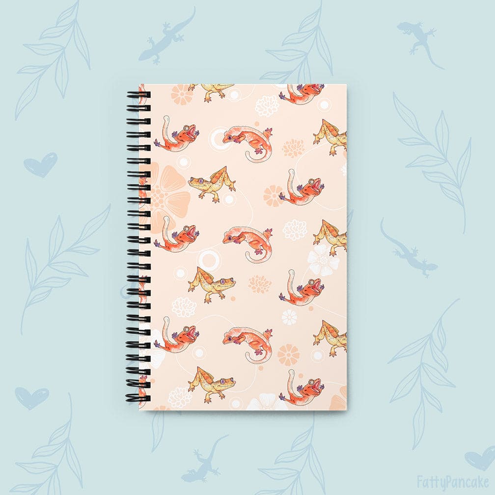 A spiral-bound notebook with a light peach cover featuring a repeating pattern of cartoon geckos in orange, yellow, and brown, set against a light blue background with faint leaf and gecko silhouettes.