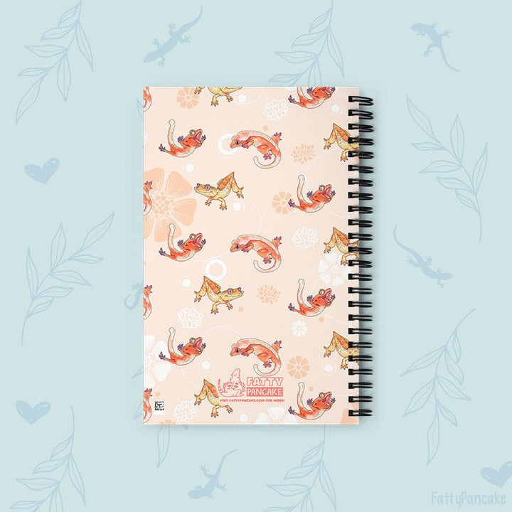 A spiral-bound notebook with a light peach cover decorated with a pattern of orange and yellow crested geckos, white flowers, and black spiral binding, set against a light blue background with subtle leaf and lizard silhouettes.