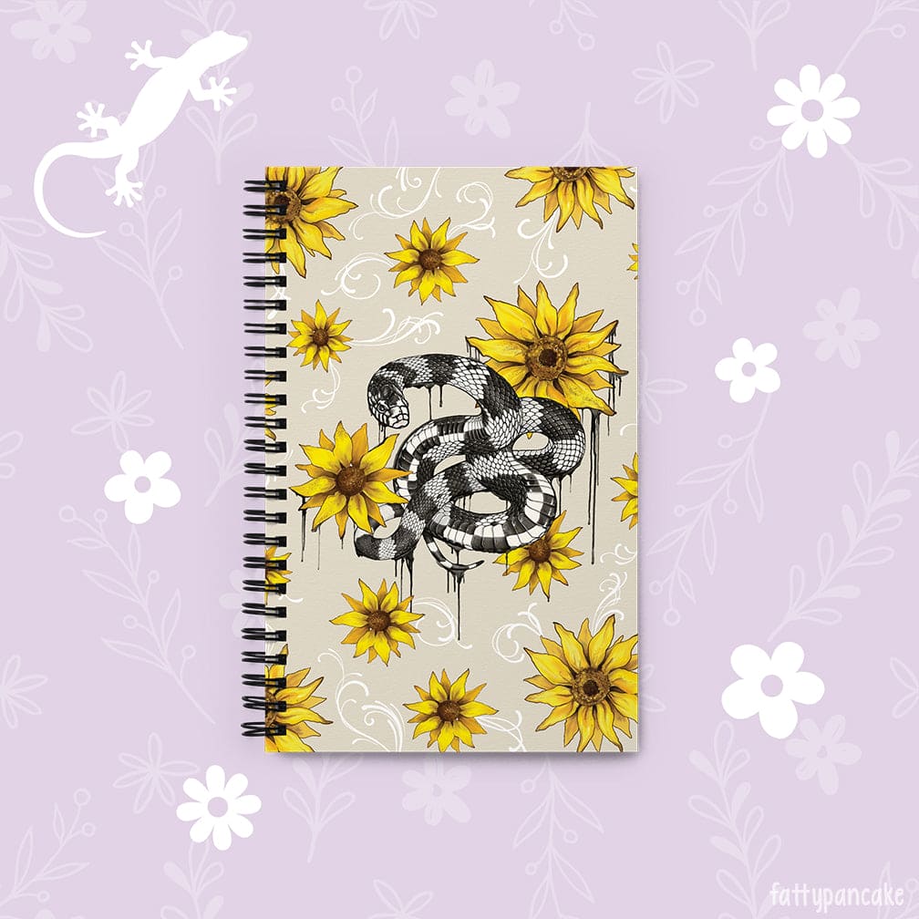 A spiral-bound notebook with a beige cover featuring a black and white snake illustration and yellow sunflower designs, displayed on a light purple background with white floral motifs.