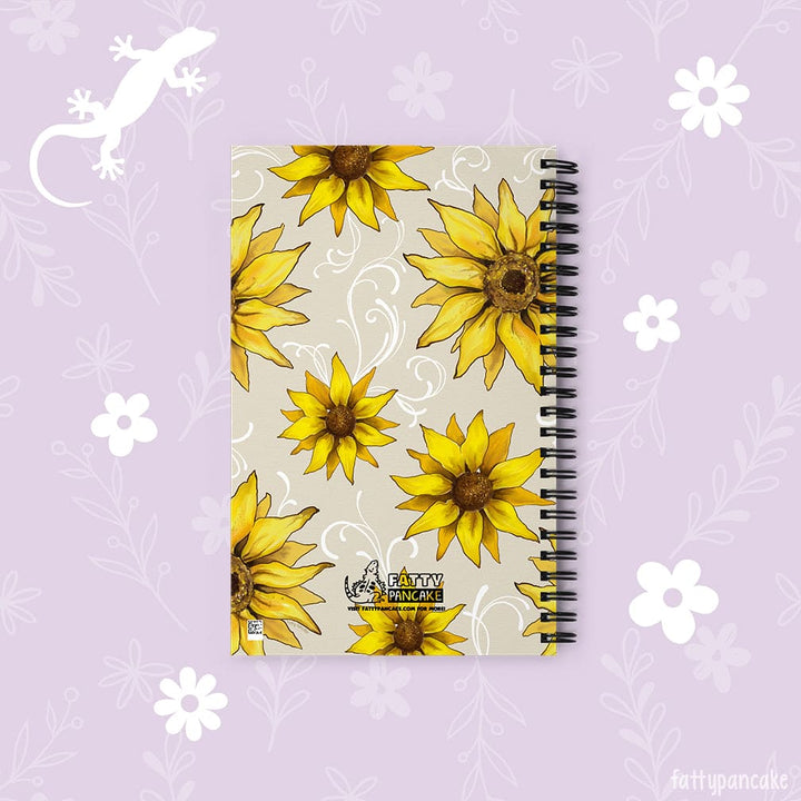 A spiral-bound notebook with a beige cover decorated with yellow sunflower illustrations and white swirl patterns, displayed on a light purple floral background.