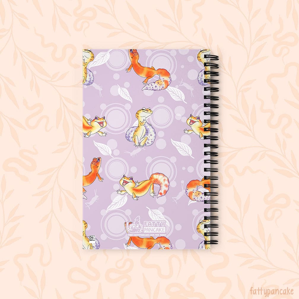 A spiral-bound notebook with a lavender cover decorated with cartoon leopard geckos, leaves, and circles, shown on a pale peach background with a faint floral pattern. This depicts the rear of the journal.
