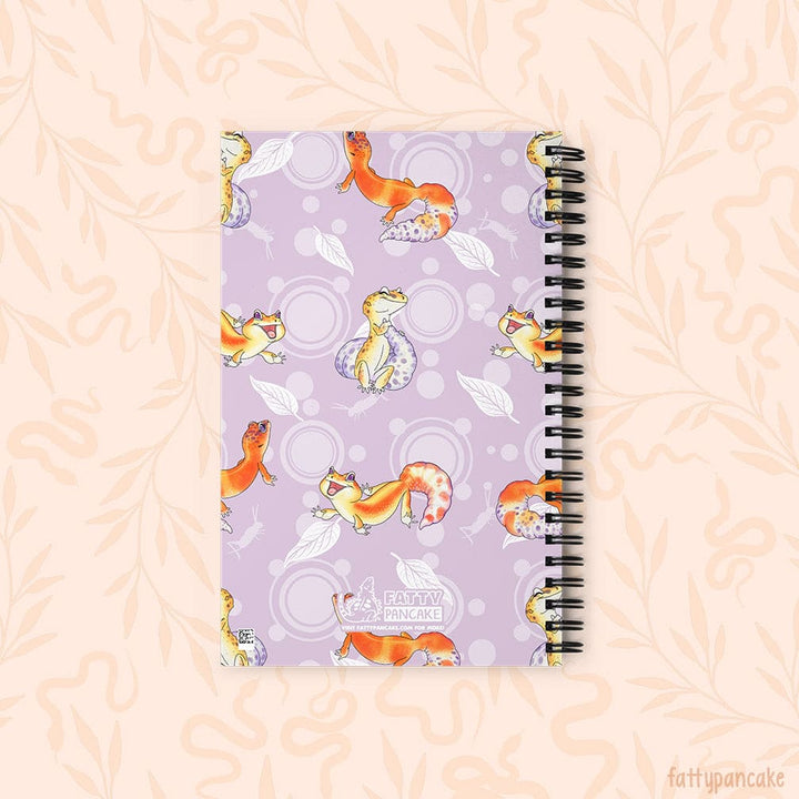 A spiral-bound notebook with a lavender cover decorated with cartoon leopard geckos, leaves, and circles, shown on a pale peach background with a faint floral pattern. This depicts the rear of the journal.