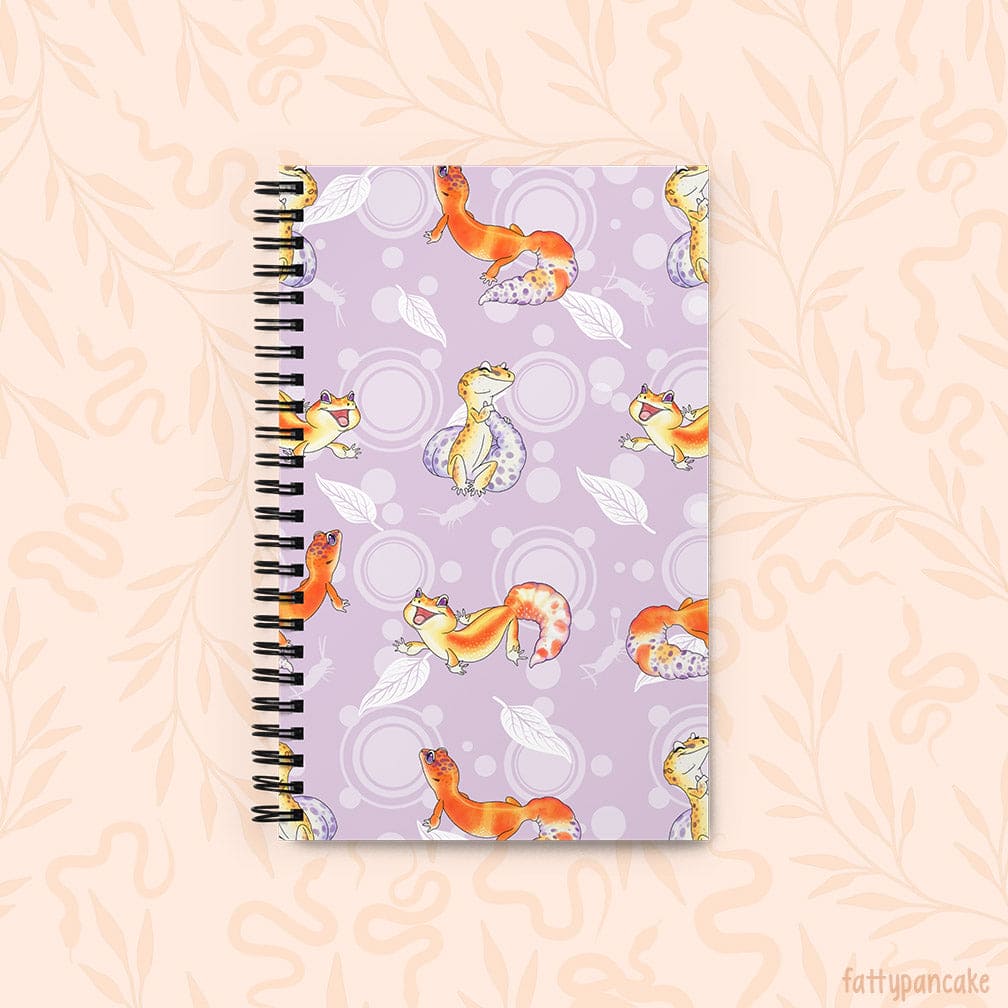 A spiral-bound notebook with a lavender cover decorated with cartoon leopard geckos, leaves, and circles, shown on a pale peach background with a faint floral pattern. This depicts the Front of the journal.