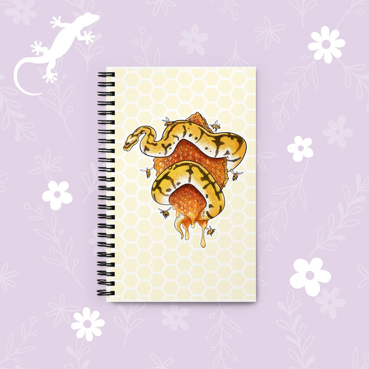 A spiral-bound notebook with a pale yellow honeycomb pattern cover, featuring an illustration of a yellow and black snake wrapped around a honeycomb structure. The notebook is displayed on a light lavender background decorated with white floral graphics and a white lizard silhouette.