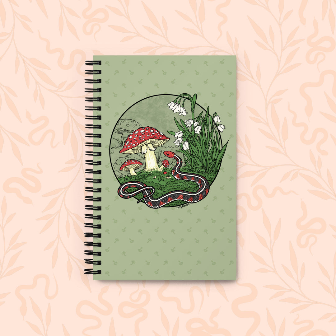 A spiral-bound notebook with a sage green cover featuring an illustration of red-and-white mushrooms, white flowers, and a red-and-black snake inside a circular frame, set against a pale peach floral background.