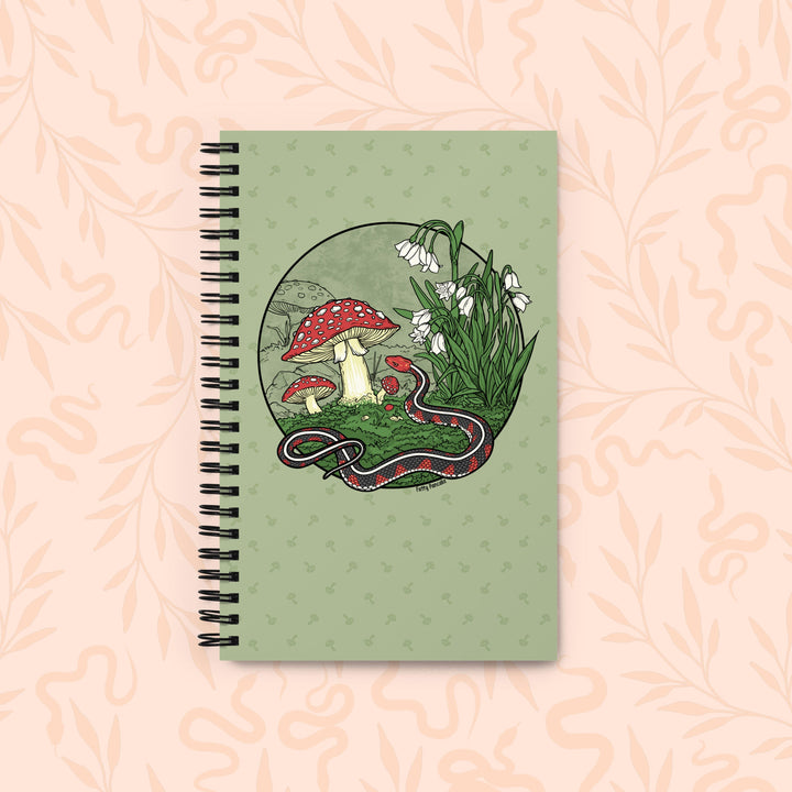 A spiral-bound notebook with a sage green cover featuring an illustration of red-and-white mushrooms, white flowers, and a red-and-black snake inside a circular frame, set against a pale peach floral background.