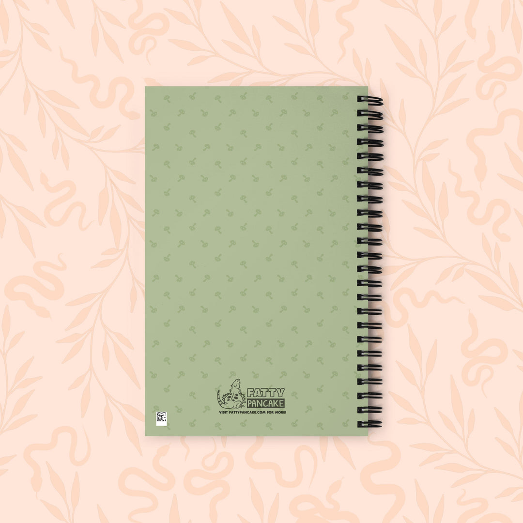 A spiral-bound notebook with a sage green cover featuring a subtle repeating pattern of mushrooms, placed on a light peach background with a faint floral design.