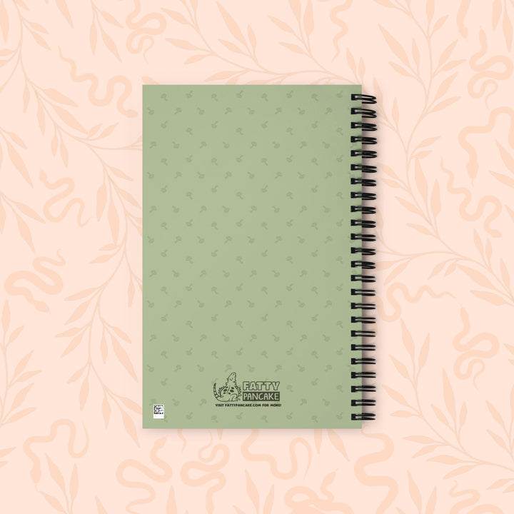 A spiral-bound notebook with a sage green cover featuring a subtle repeating pattern of mushrooms, placed on a light peach background with a faint floral design.