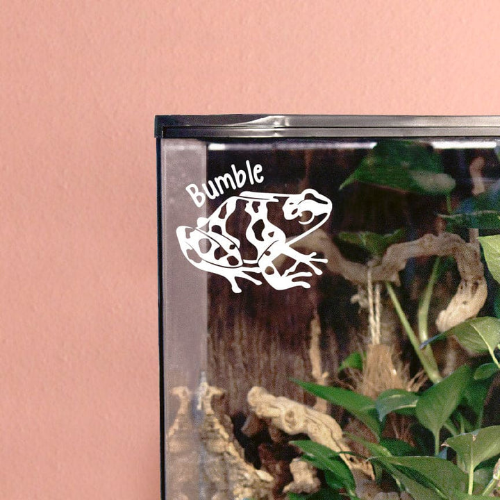 Photo depicts a white vinyl decal sticking on the top corner of a glass vertical enclosure. The decal design shows a Bumblebee Dart Frog, it's strong adhesive making it a permanent fixture to it's surface, highlighting the brilliant matte white color and smooth finish of the material.