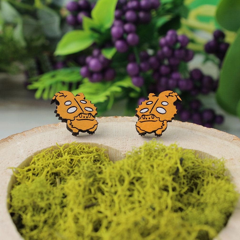 A pair of wooden post earrings featuring a cartoon bearded dragon. The earrings are displayed on a bed of green moss with purple decor in the background.