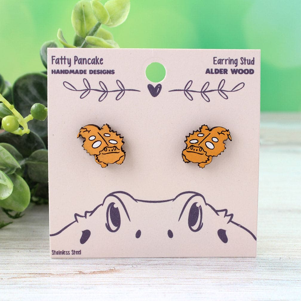 Hand painted wooden stud earrings with an orange cartoon smiling bearded dragon with white spots, mounted on a card with the brand name &