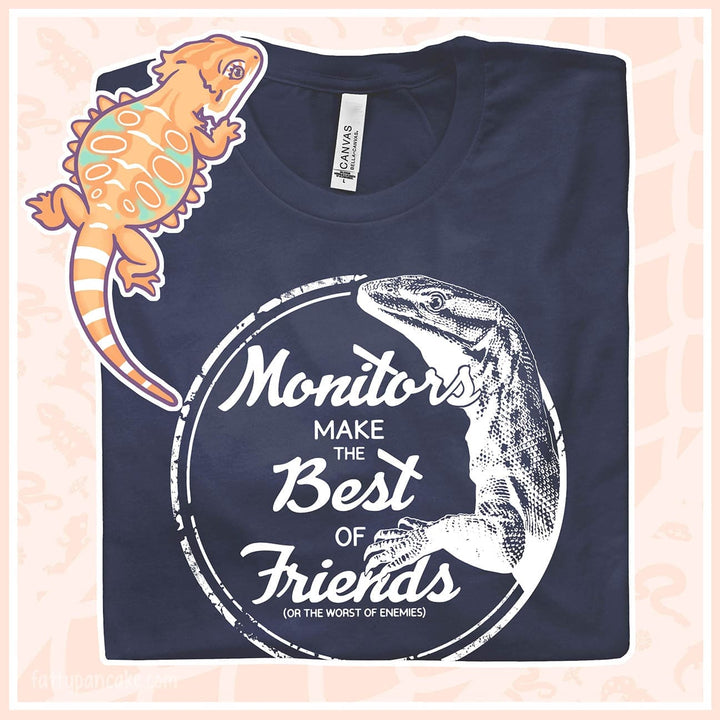 Navy blue image of a Bella Canvas tee with a large white illustration of an ackie monitor framed by a circle and the text, "Monitors make the Best of Friends" (or the worst of enemies. The shirt is a top down image with a cartoon bearded dragon resting on the surface.