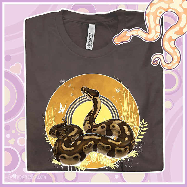 Image shows folded brown colored crew neck tee with a ball python resting atop of a circle with warm amber colors of fall with simple yellow leaf designs.