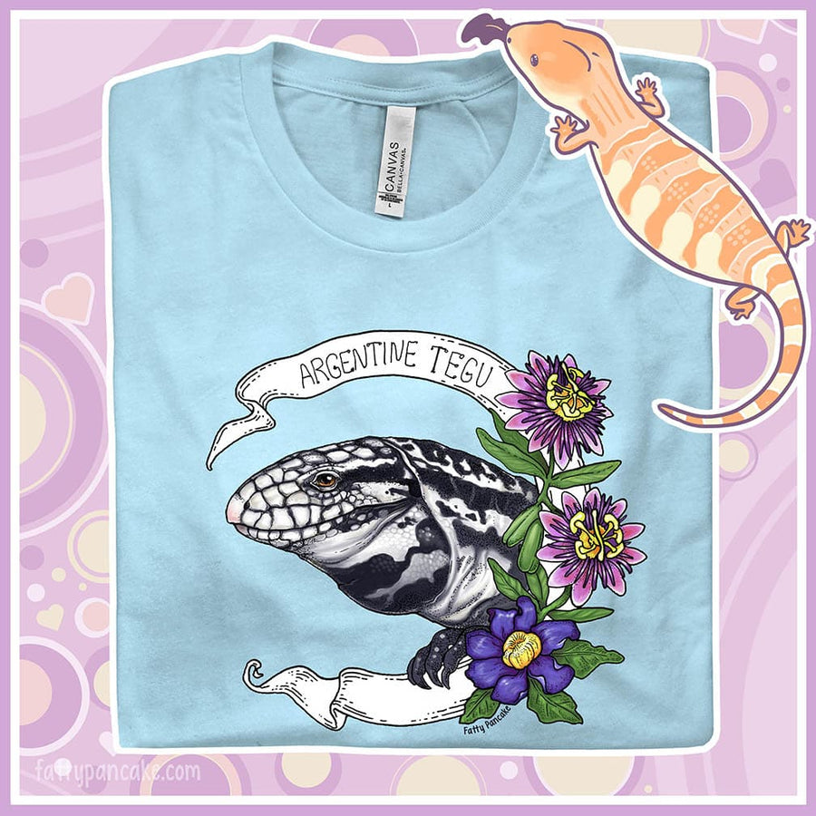 Image shows a folded crew neck tee to show an enlarged graphic of an Argentine Tegu lizard. Purple and pink flowers are on the right of the graphic and a white banner encircles the reptile.