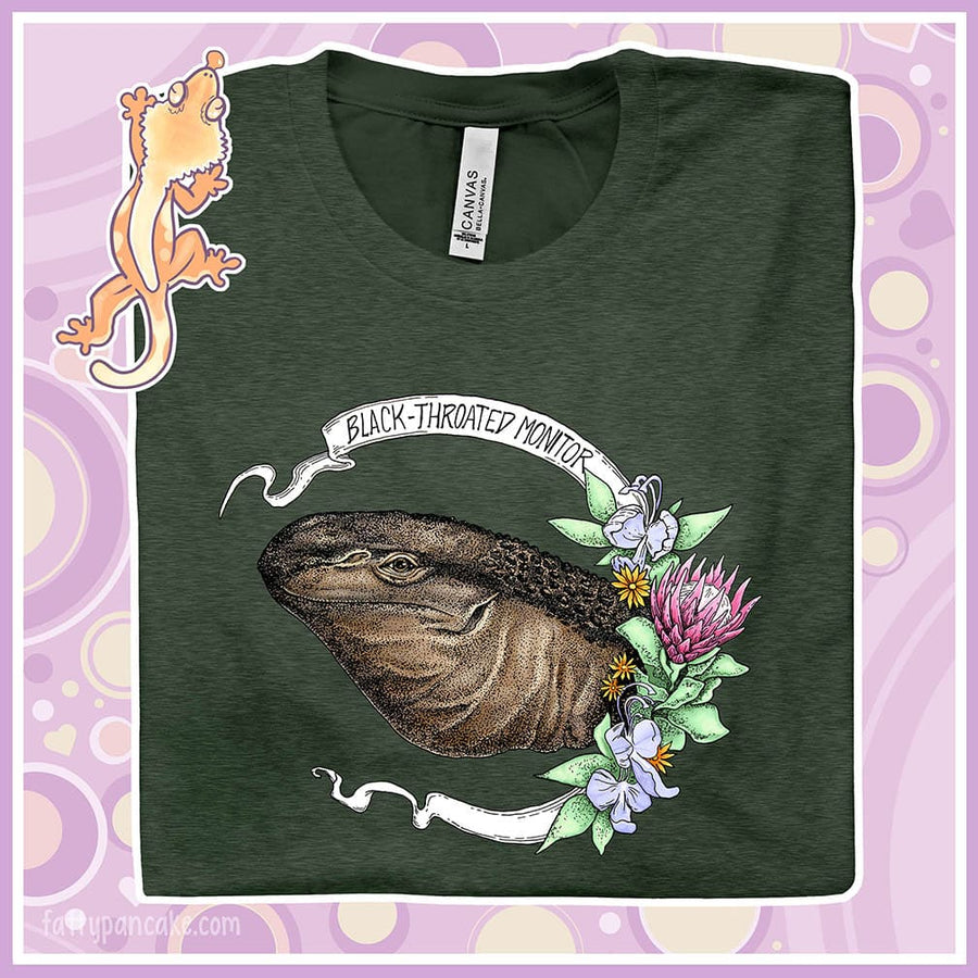 Folded green colored crew neck tee of a black throated monitor encircled by a banner that reads the species name and with rendered blue and red flowers next to it.