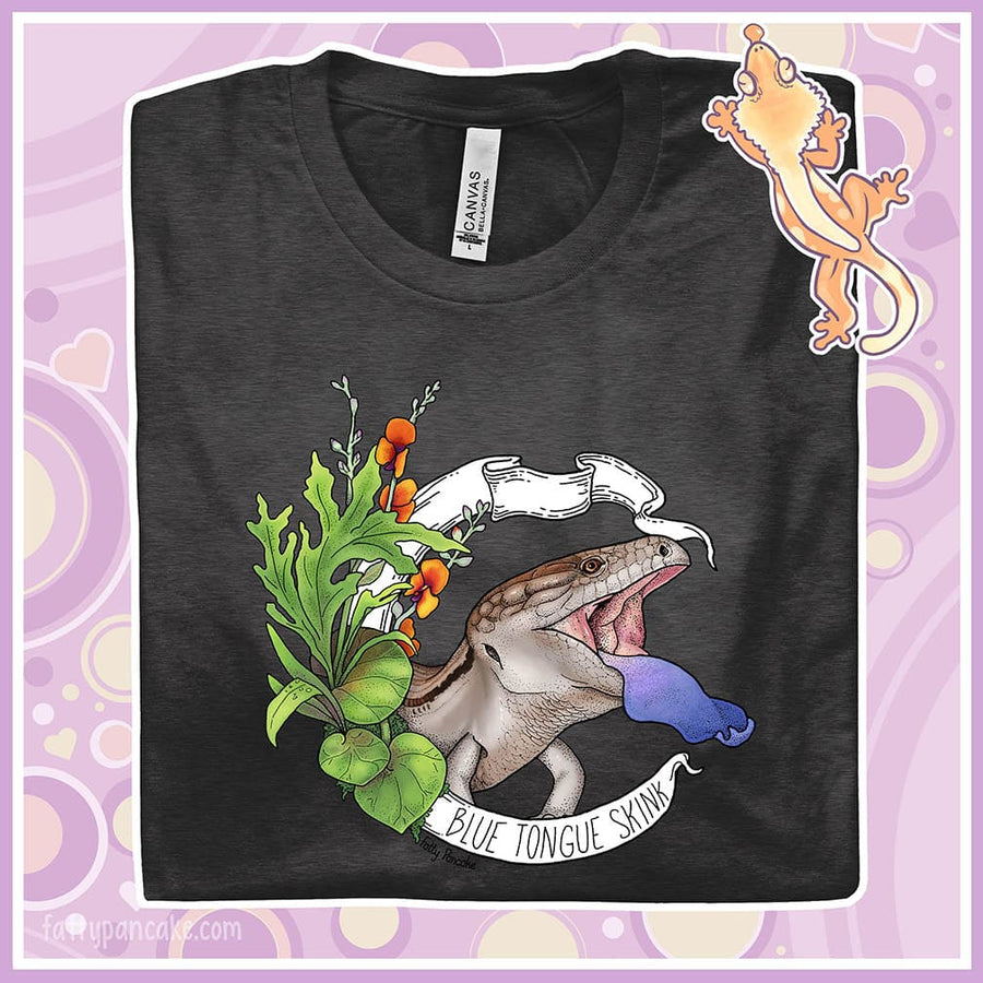 Folded grey tee with a hand drawn design of a blue tongue skink flashing it's blue tongue in warning. Framed with greenery and a white banner that reads, "Blue Tongue Skink".